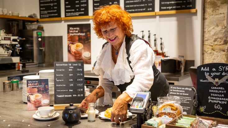 A white woman with bright red hair is standing behind a café counter. The wall behind her is filled with black chalk menu boards and the counter is filled with cakes. In front of her is a black teapot and cup and saucer.